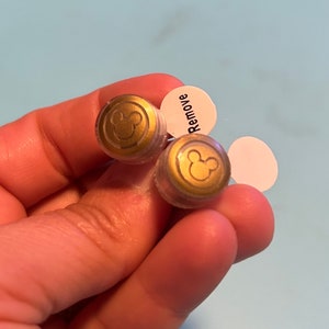 May include: Two clear plastic earring backs with a gold Mickey Mouse silhouette on each. The backs have a white sticker with the word "Remove" on one of them.