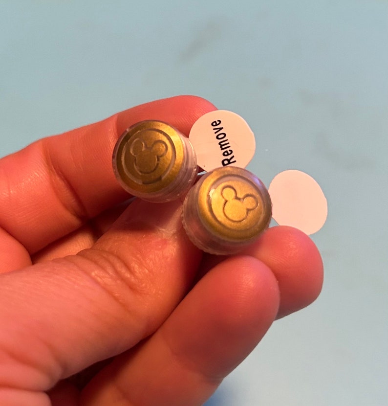 以下が含まれることがあります： 透明なプラスチック製のイヤリングバックが2つ。それぞれに金色のミッキーマウスのシルエットが描かれています。バックの1つには、「Remove」と書かれた白いステッカーが付いています。