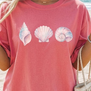 Pode incluir: Camiseta coral com três ilustrações de conchas em cores pastel suaves. A camisa tem um corte folgado e mangas curtas. A palavra "Watermelon" está impressa no canto superior esquerdo.