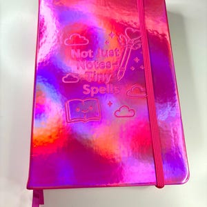 May include: A pink, holographic journal with the text "Not Just Notes - Tiny Spells." The cover features embossed cloud and star designs, a wand, and a smiling book. It has a pink elastic band closure and a ribbon bookmark.