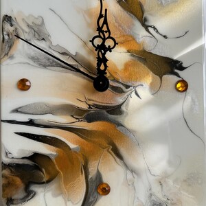 May include: Square wall clock with a white background and an abstract design in gold, black, and gray. The clock has black hands and amber-colored markers. The clock face has a glossy finish.