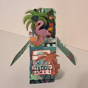 May include: A tropical-themed pop-up card featuring a pink glitter flamingo, palm trees, and a parrot. The card includes the phrase "Be HAPPY" on a wooden sign. The background has a tropical leaf pattern.