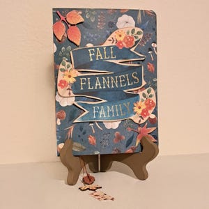 May include: A rectangular card featuring a dark blue background with a pattern of autumn leaves, mushrooms, and flowers. A central blue ribbon banner displays the gold text FALL FLANNELS FAMILY. The card is displayed on a brown wooden stand.