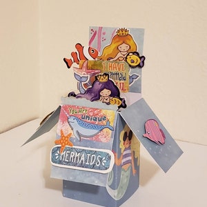May include: A blue and white pop-up card with a mermaid theme. The card features a mermaid with a crown, a narwhal, a starfish, and the text "You are unique" and "Mermaids".