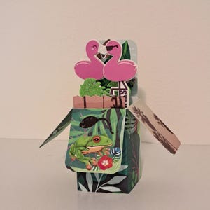 May include: A decorative pop-up card featuring a tropical theme. The card showcases two pink flamingo cutouts, one wearing sunglasses. The front panel displays a green frog, a black insect, and tropical foliage. The card has a box shape.
