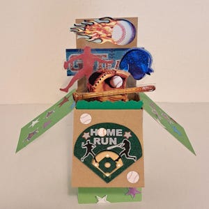 May include: A three-dimensional pop-up card featuring baseball imagery. The front displays a green diamond with the text HOME RUN and silhouettes of players. The top section includes a baseball with flames, a glove, a bat, and a blue helmet.