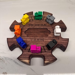 May include: A dark wood Mexican Train hub with eight slots, each holding a small plastic train figurine in yellow, green, gray, white, black, pink, blue, and orange. The center of the wooden hub is engraved with the text MEXICAN TRAIN.
