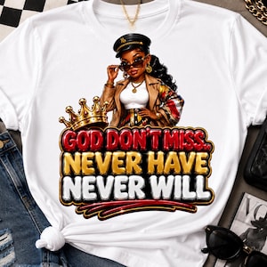 May include: A white t-shirt featuring a graphic of a person wearing a tan jacket, black cap, and sunglasses, positioned above a gold crown and the text "GOD DON'T MISS.. NEVER HAVE NEVER WILL" in bold, layered red, gold, and white lettering.