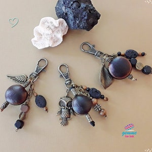 Könnte beinhalten: Drei Schlüsselanhänger mit braunen Samenkapseln, Metallanhängern und schwarzen Lavasteinen. Ein Schlüsselanhänger hat einen kleinen Eulenanhänger. Die Schlüsselanhänger befinden sich auf einer hellbraunen Oberfläche.