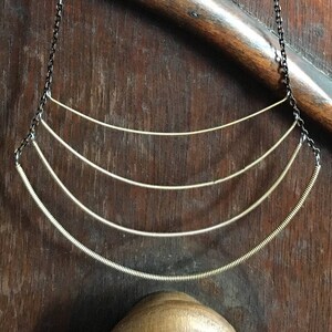 Op de afbeelding: Een zilveren ketting met meerdere lagen gebogen draad, hangend aan een zwarte ketting.