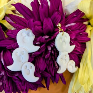 Może przedstawiać: Para białych kolczyków w kształcie ducha z złotymi haczykami. Kolczyki są wykonane z polimerowej gliny i są zabawnym i upiornym dodatkiem na Halloween.