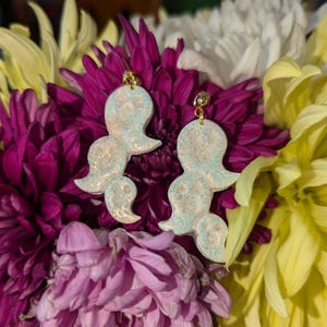 Op de afbeelding: Een paar witte, iriserende oorbellen in de vorm van een spook met gouden haakjes. De oorbellen zijn gemaakt van polymer clay en hebben een glinsterende afwerking.