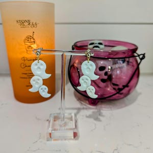 Pode incluir: Brincos brancos em forma de fantasma com detalhes dourados. Os brincos est&atilde;o pendurados em um expositor transparente. Os brincos est&atilde;o na frente de um caldeir&atilde;o rosa com um design de caveira.