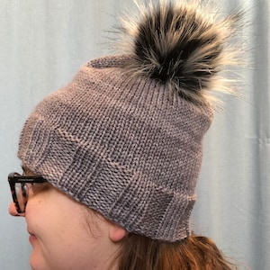 Puede incluir: Un gorro de punto gris con un pompón de piel sintética negro y blanco.