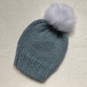May include: A knitted beanie in a muted teal color, topped with a large, fluffy white pom-pom. The hat is made of a thick, textured yarn, perfect for cold weather. The pom-pom adds a playful touch to the winter accessory.