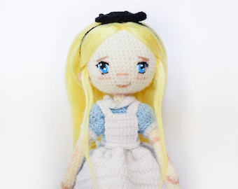 May include: A crocheted doll with blonde hair, a blue dress, a white apron, and black shoes. The doll is wearing a black hat. The doll is standing on a white background.