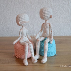 May include: Two white crocheted dolls sitting on balls of yarn. One doll is sitting on a pink ball of yarn and the other doll is sitting on a blue ball of yarn.