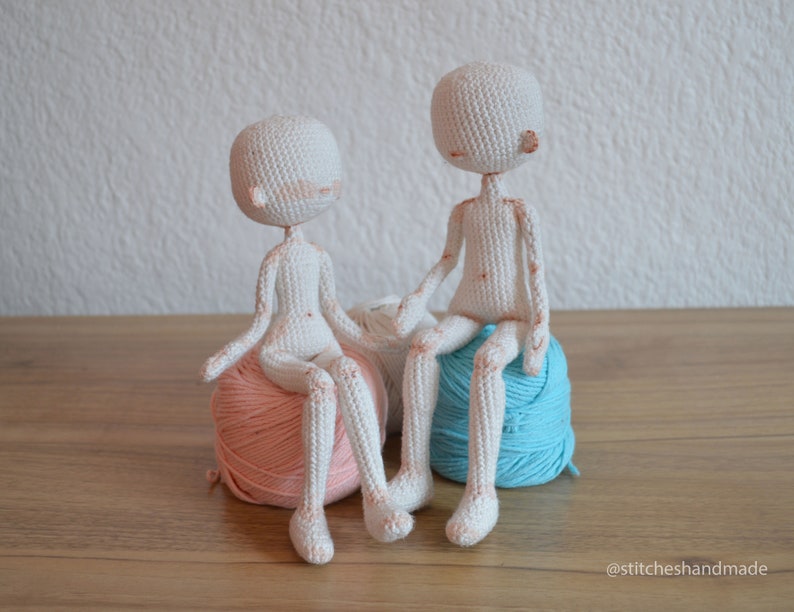 May include: Two white crocheted dolls sitting on balls of yarn. One doll is sitting on a pink ball of yarn and the other doll is sitting on a blue ball of yarn.