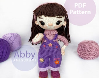 May include: Crochet amigurumi doll pattern for a girl named Abby wearing a purple overall dress with a pink sweater and brown hair. The pattern is available as a PDF download. The image also includes purple and pink yarn balls.