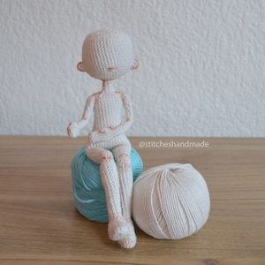 May include: A white crocheted doll sitting on a blue yarn ball with a white yarn ball beside it. The doll has a simple face with no eyes or mouth. The doll is sitting with its legs crossed and its arms folded in front of it. The text "@stitcheshandmade" is visible in the lower right corner of the image.