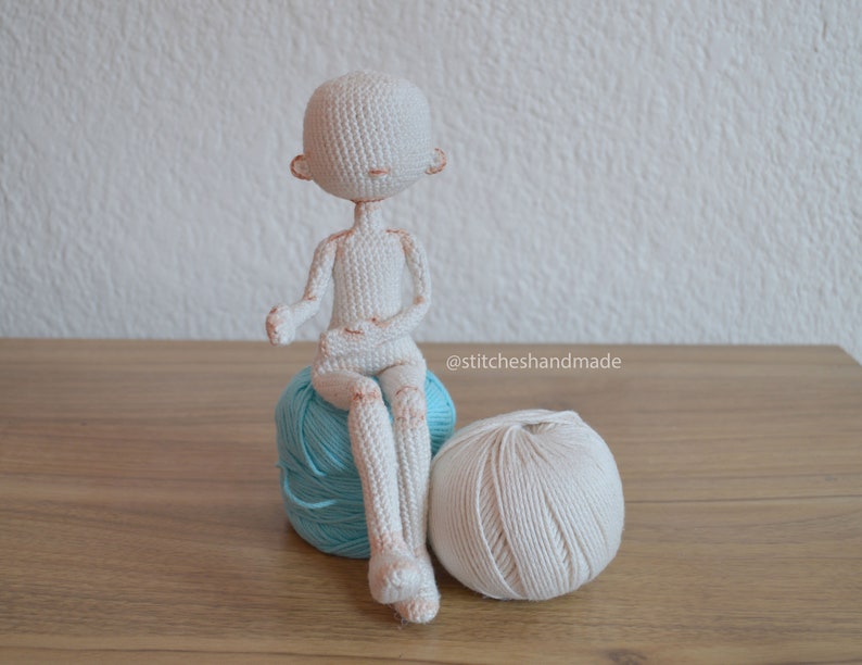 May include: A white crocheted doll sitting on a blue yarn ball with a white yarn ball beside it. The doll has a simple face with no eyes or mouth. The doll is sitting with its legs crossed and its arms folded in front of it. The text "@stitcheshandmade" is visible in the lower right corner of the image.