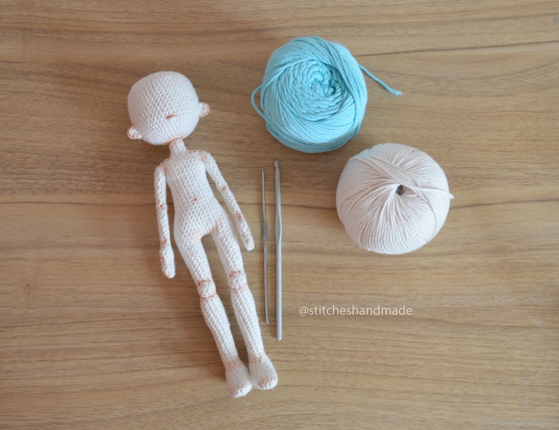 May include: A white crocheted doll with a light blue and a beige ball of yarn and two crochet hooks on a wooden surface. The text '@stitcheshandmade' is visible in the bottom right corner of the image.
