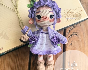 May include: Crocheted doll with purple flower crown, purple dress, and a purple cardigan. The doll is holding a small brown bag. The doll is sitting on a wooden surface. The text "Lyla PDF Pattern" is visible in the image.