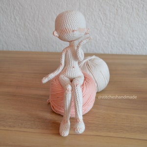 May include: A white crocheted doll with a pink blush sitting on a pink and white yarn ball. The doll has a simple design and is sitting with its legs crossed. The yarn ball is on a wooden surface.
