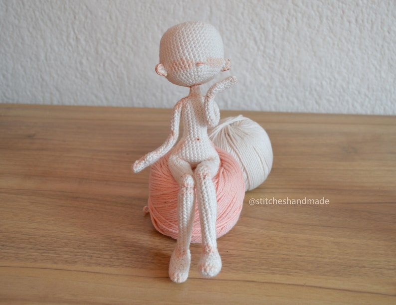 May include: A white crocheted doll with a pink blush sitting on a pink and white yarn ball. The doll has a simple design and is sitting with its legs crossed. The yarn ball is on a wooden surface.