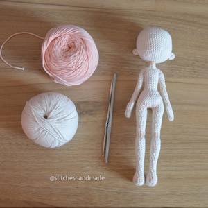 May include: A white crocheted doll with a pink yarn ball and a white yarn ball. The doll is missing its arms and legs. There is a crochet hook next to the yarn balls.