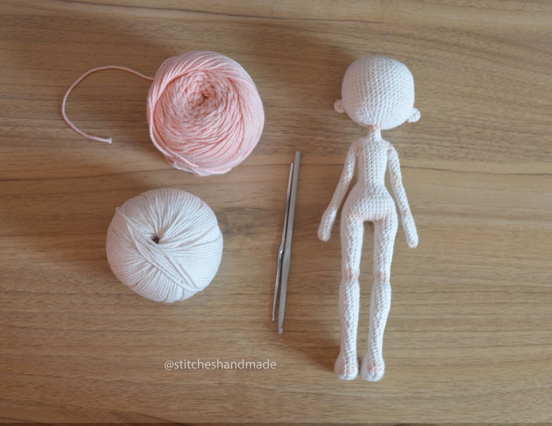 May include: A white crocheted doll with a pink yarn ball and a white yarn ball. The doll is missing its arms and legs. There is a crochet hook next to the yarn balls.