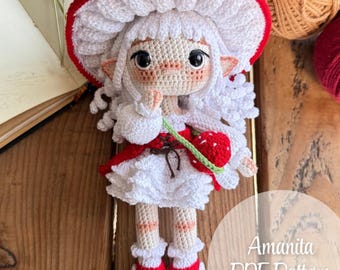 May include: A handmade crochet doll with a mushroom hat, white dress, and red accents. The doll has a strawberry accessory and is labeled "Amanita PDF Pattern" with the Instagram handle "@stitcheshandmade".