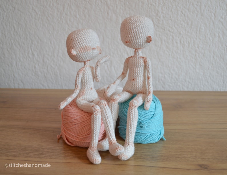 May include: Two crocheted dolls sitting on balls of yarn. The doll on the left is sitting on a peach-colored ball of yarn, and the doll on the right is sitting on a light blue ball of yarn.