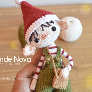 Puede incluir: Una muñeca de elfo de crochet con un gorro rojo y blanco, un mono verde y una bolsa marrón. La muñeca tiene el pelo de lana marrón y ojos negros.