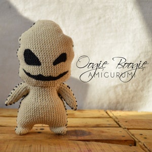 Puede incluir: Un juguete amigurumi de crochet de Oogie Boogie de la película Pesadilla antes de Navidad. El juguete es beige con ojos negros y una boca negra. El juguete está de pie sobre una superficie de madera. El texto "Oogie Boogie Amigurumi" está escrito en cursiva negra sobre un fondo blanco.