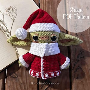 Puede incluir: Muñeco Grogu de crochet vestido con un traje de Papá Noel rojo y blanco con un gorro blanco de crochet. El muñeco está sentado sobre una superficie de madera con un libro y una rama de flores blancas detrás. El texto "Grogu PDF Pattern" es visible en la esquina superior derecha de la imagen.