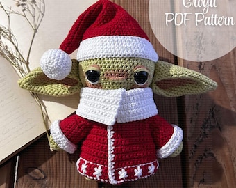 May include: Crocheted Grogu doll dressed in a red and white Santa suit with a white crocheted hat. The doll is sitting on a wooden surface with a book and a sprig of white flowers behind it. The text "Grogu PDF Pattern" is visible in the upper right corner of the image.