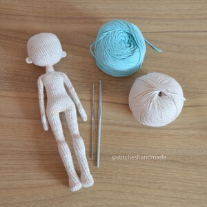 May include: A crocheted doll with a white yarn body and a light blue yarn ball and a beige yarn ball. The doll is on a wooden surface with two crochet needles.
