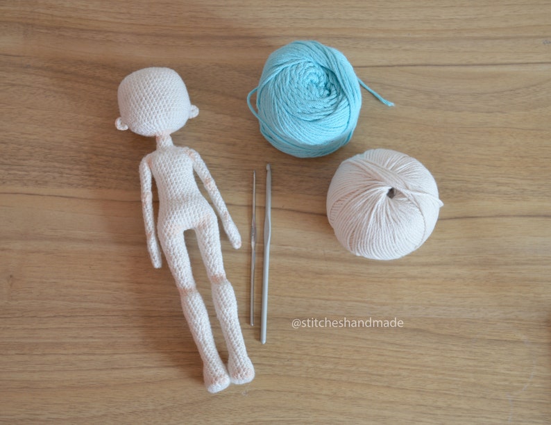 May include: A crocheted doll with a white yarn body and a light blue yarn ball and a beige yarn ball. The doll is on a wooden surface with two crochet needles.