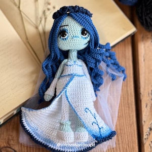 May include: A handmade crochet doll in the style of the Corpse Bride. The doll has blue hair, a pale green face, and wears a white dress with blue accents. The doll is sitting on a wooden surface.