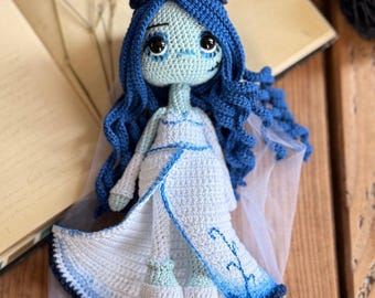 May include: A handmade crochet doll in the style of the Corpse Bride. The doll has blue hair, a pale green face, and wears a white dress with blue accents. The doll is sitting on a wooden surface.