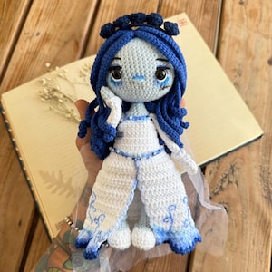 May include: A crocheted doll with blue hair and a white dress. The doll has a pale blue face with large eyes and a small mouth. The doll is wearing a white dress with blue accents and a veil.