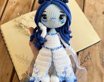 May include: A crocheted doll with blue hair and a white dress. The doll has a pale blue face with large eyes and a small mouth. The doll is wearing a white dress with blue accents and a veil.