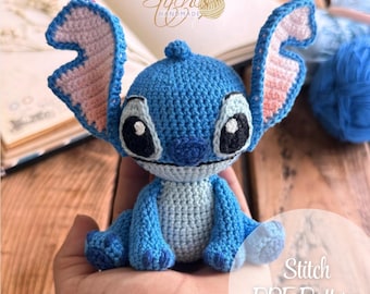 May include: A blue crocheted Stitch character from the Disney movie Lilo & Stitch. The character has large ears, big eyes, and a small mouth. Stitch is sitting on a hand.