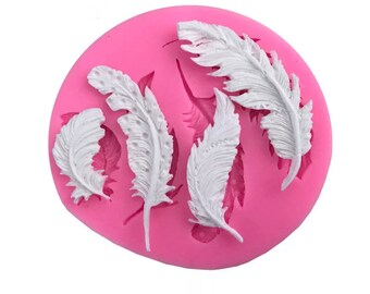 May include: A pink silicone mold featuring five white feather designs. The feathers are intricately detailed, with visible veins and textures. The mold is round and likely used for crafting or baking decorative elements.