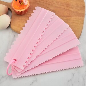 May include: A collection of pink plastic cake decorating scrapers with different edge designs. The scrapers are fanned out, displaying various textured edges. A pink ring holds the scrapers together. A cupcake and eggs are in the background.