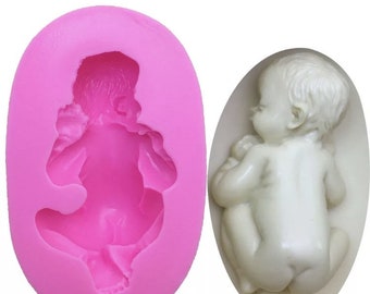 May include: Pink silicone mold for making a baby figurine. The mold is shaped like a sleeping baby with arms and legs curled up. The mold is empty and ready to be filled with fondant or other edible material.