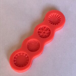 May include: A red silicone mold with four different designs. The mold is shaped like a long, thin rectangle with rounded edges. The designs are all circular and feature intricate patterns.