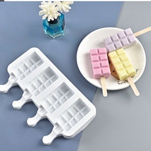 May include: White silicone popsicle mold with four slots. Each slot has a grid pattern and a handle. The mold is on a gray surface. Three popsicles are on a white plate with wooden sticks. The popsicles are pink, white, and purple.