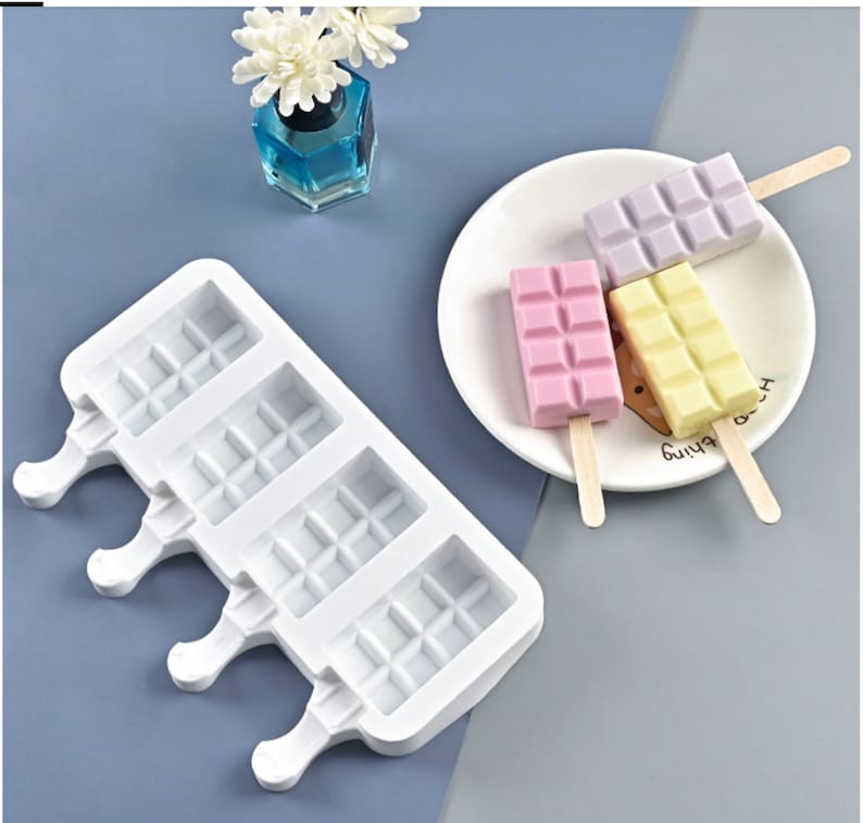 May include: White silicone popsicle mold with four slots. Each slot has a grid pattern and a handle. The mold is on a gray surface. Three popsicles are on a white plate with wooden sticks. The popsicles are pink, white, and purple.
